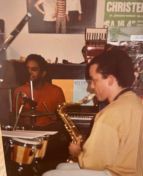 Farbfoto, frühe 1990er Jahre: Ronnie Burrage am Schlagzeug und John Purcell am Saxofon bei einer Probe im Heimstudio in Hernstein; an der Wand das Plakat des Trios Pepl, Joos, Christensen und ein Akkordeon. Links Ronnie Burrage am Schlagzeug, rechts John Purcell am Saxofon.
