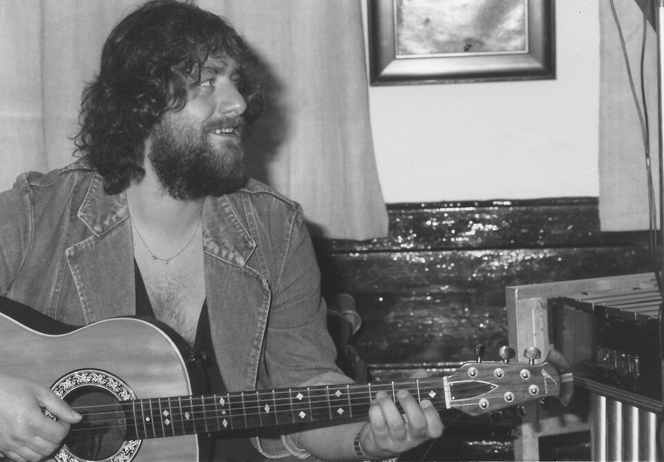 Schwarzweißfoto aus dem Familienarchiv, September 1979: Harry Pepl mit lockigem Haar und Vollbart, in Jeansjacke, spielt eine Akustikgitarre und blickt lächelnd zur Seite, rechts ein Gerät mit Metallstäben.