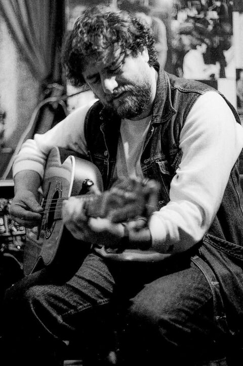 Schwarzweißfoto: Harry Pepl in Jeansweste spielt sitzend eine akustische Gitarre mit Tonabnehmer, der Blick ist auf die Saiten gerichtet, die Greifhand verwischt in der Bewegung.