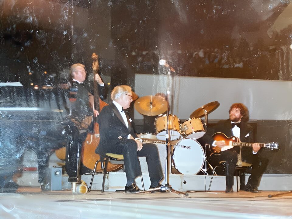 Stark beschädigter Farbabzug einer Bühnenszene: ein Bassist, ein sitzender Mann im Smoking, ein Schlagzeug und rechts ein Gitarrist im Scheinwerferlicht. Von links nach rechts: Peter Witte am Kontrabass, Benny Goodman sitzend im Smoking und Harry Pepl mit der Gitarre; der Schlagzeuger ist verdeckt.