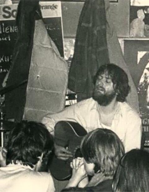 Sepiafarbener Abzug: der junge Harry Pepl mit Vollbart spielt in einem kleinen Club Akustikgitarre, an der Wand Plakate, im Vordergrund die Köpfe des Publikums. Aufnahmejahr und Ort sind nicht überliefert.