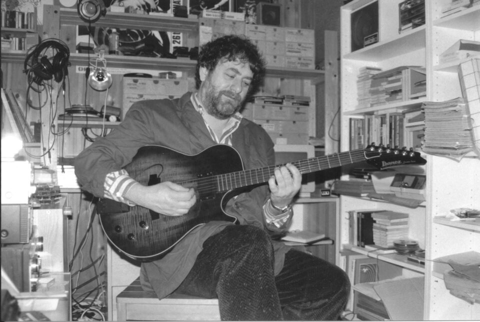 Schwarzweißfoto: Harry Pepl sitzt in seinem Heimstudio mit einer Halbresonanz-E-Gitarre der Marke Ibanez und blickt lächelnd auf das Griffbrett; hinter ihm Regale mit Archivkästen, ein Kopfhörer am Haken, links ein Tonbandgerät.
