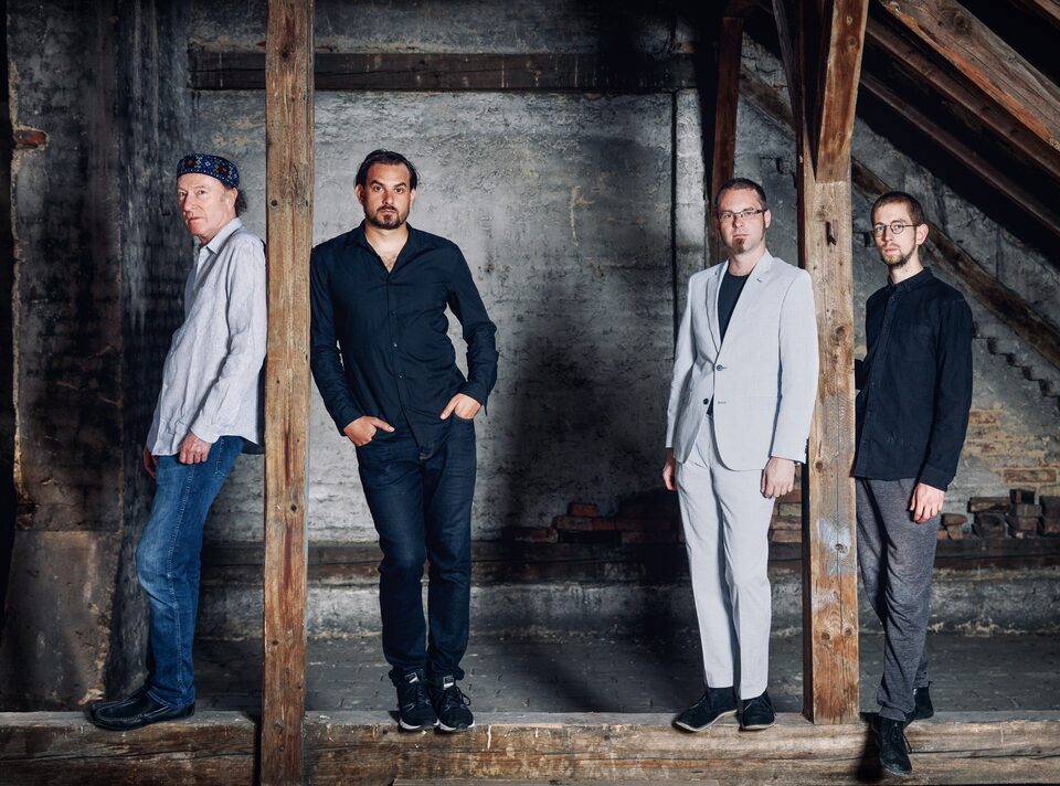 Farbfoto: vier Männer in dunkler Kleidung stehen zwischen den Balken eines Dachbodens vor einer Ziegelwand, der linke im Profil, die anderen blicken in die Kamera. Von links: Andreas Schreiber (Violine), Martin Bayer (Gitarre), Clemens Salesny (Saxophon) und Valentin Duit (Schlagzeug).