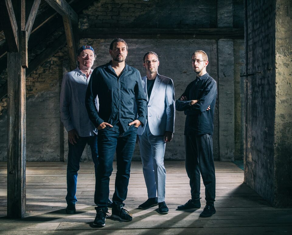 Farbfoto: vier Männer stehen in einem Dachboden mit alten Holzbalken und Ziegelwänden, dunkle Kleidung, ernste Haltung, Streiflicht von links. Von links: Andreas Schreiber (Violine), Martin Bayer (Gitarre), Clemens Salesny (Saxophon) und Valentin Duit (Schlagzeug).