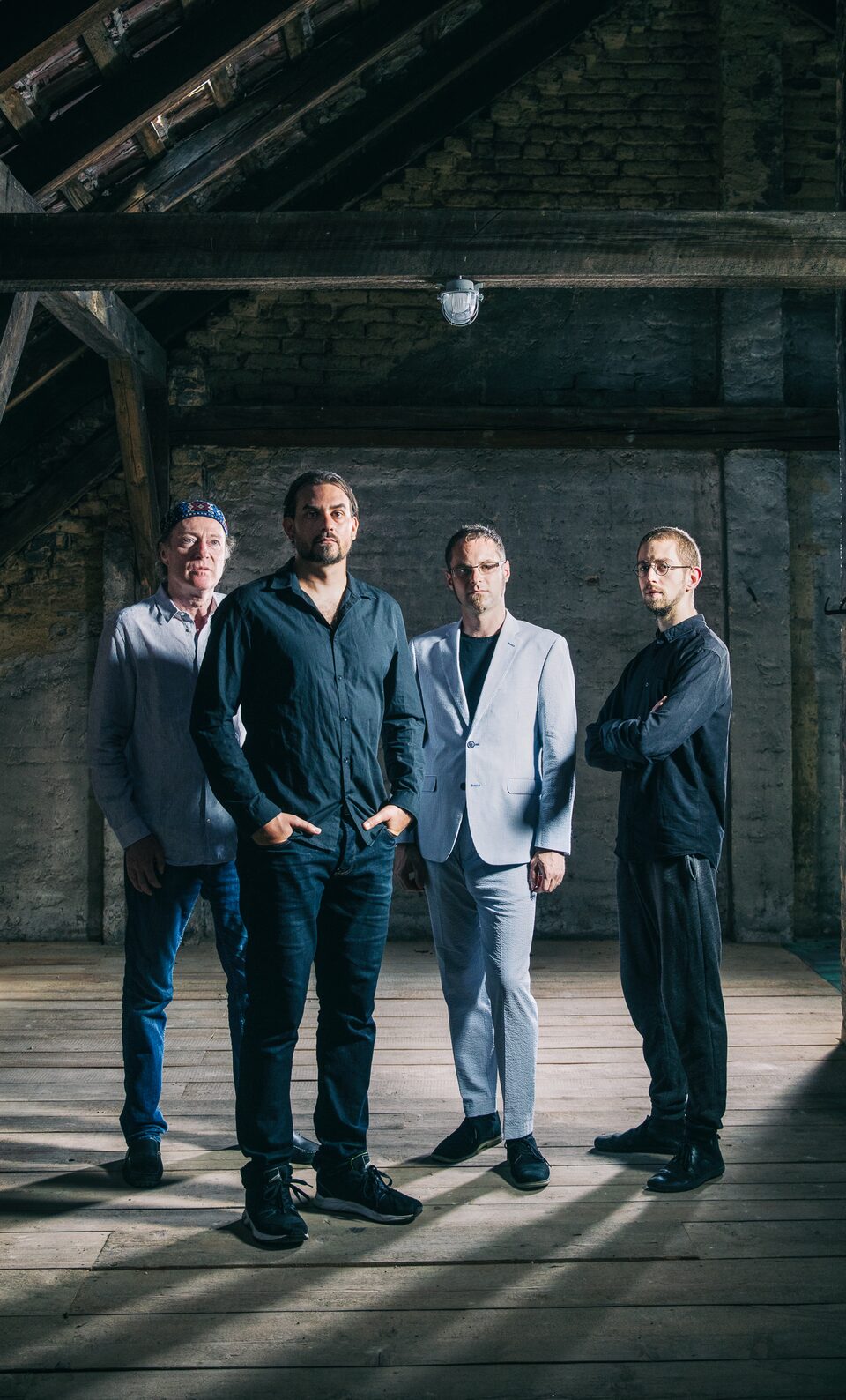 Farbfoto im Hochformat: vier Männer stehen in einem Dachboden unter Holzbalken, im Hintergrund eine Ziegelwand und eine Lampe. Von links: Andreas Schreiber (Violine), Martin Bayer (Gitarre), Clemens Salesny (Saxophon) und Valentin Duit (Schlagzeug).