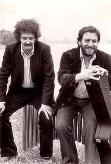 Werner Pirchner (links) und Harry Pepl sitzen lächelnd im Gras an der Alten Donau in Wien, Pirchner vor den Resonanzröhren eines Vibraphons, Pepl mit dem Gitarrenkoffer; aufgenommen in den 1970er Jahren (Angabe Daniel Pepl). Kleiner Abzug aus dem Nachlass.