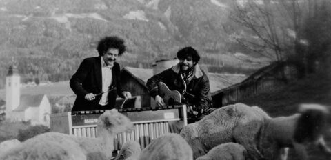 Im Freien vor einer Bergkulisse mit Dorfkirche, vermutlich in Tirol: Werner Pirchner spielt Vibraphon, Harry Pepl Gitarre, im Vordergrund drängt sich eine Schafherde. 1970er Jahre. Fotografiert hat laut Daniel Pepl Werner Pirchners Bruder. Links Werner Pirchner am Vibraphon, rechts Harry Pepl mit der Gitarre.
