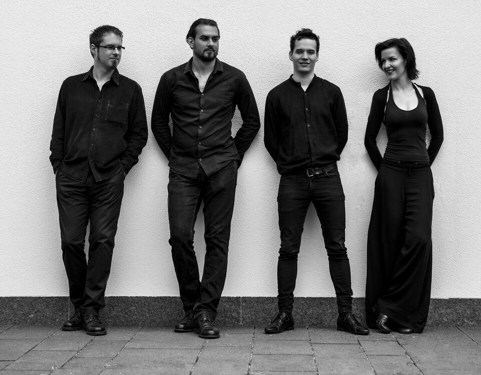 Black-and-white group picture: three male musicians and one female musician stand side by side in front of a white wall on paving stones and look into the camera. From left: Clemens Salesny, Martin Bayer, Peter Primus Frosch and Agnes Heginger.